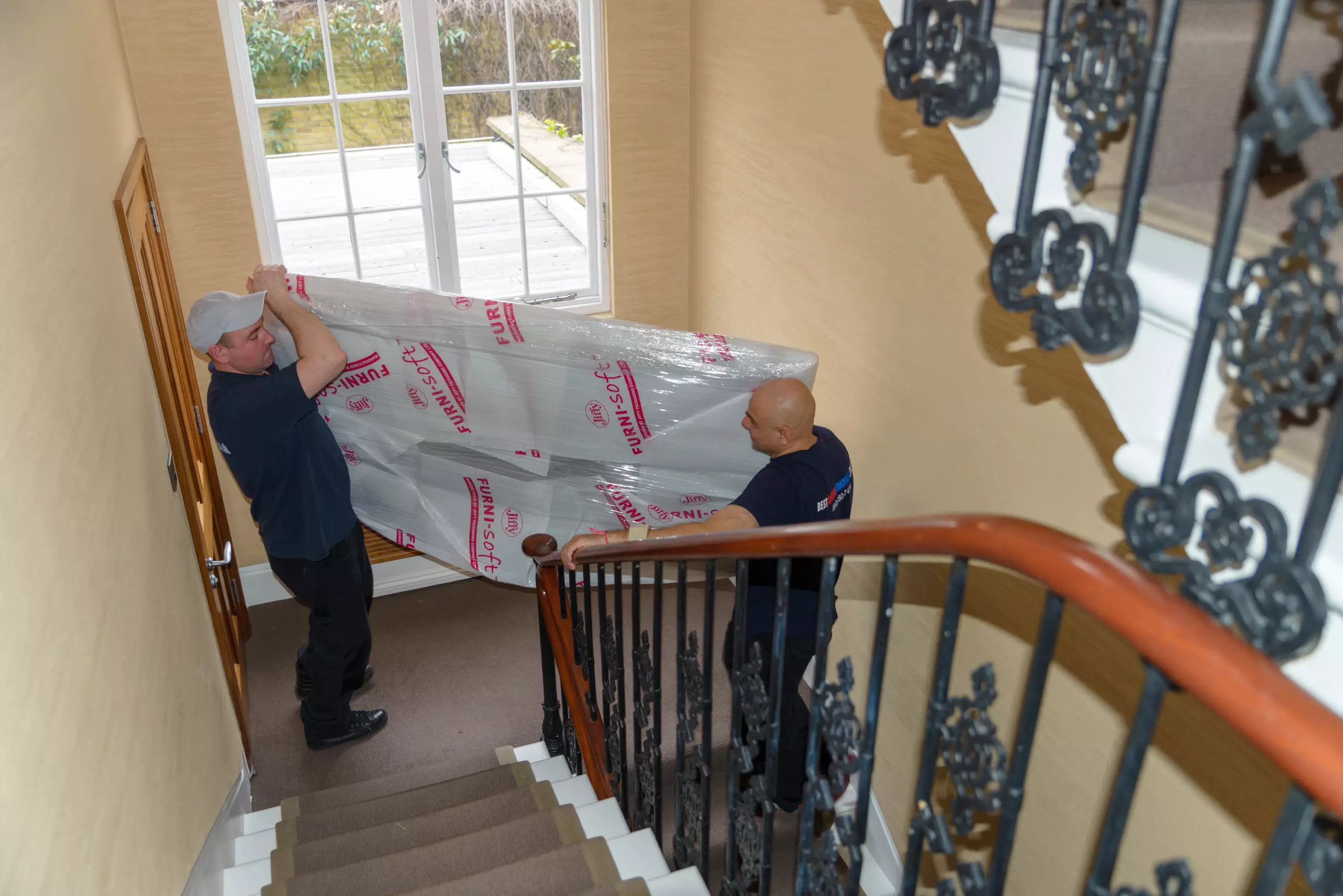 Two movers carrying a fully wrapped 77-inch TV down a staircase, packed securely for transport