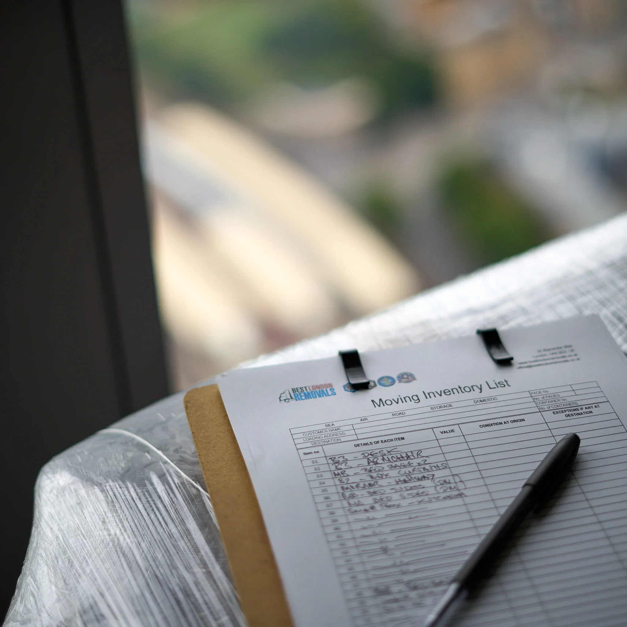Best London Removals moving inventory checklist on a clipboard with a pen placed on top near a window with a blurred city view