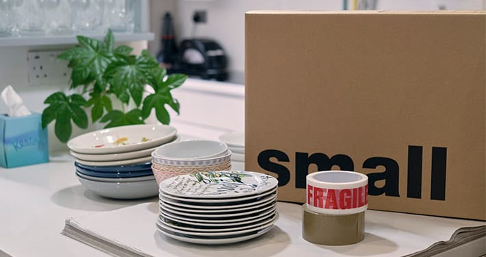 Plates and fragile packing tape next to a small labelled moving box on a kitchen counter.