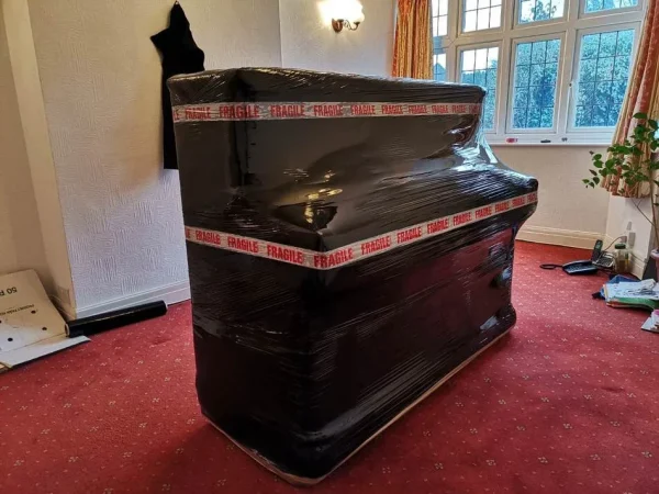 Upright piano securely wrapped in protective Furni-Guard, moving blanket and a layer of black film with ‘fragile’ tape for moving